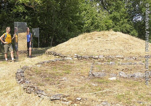 An der Grabhügelanlage stehen zwei Wanderer in sommerlicher Kleidung an einer Stele und bestaunen die Hügelanlage. Hinter ihnen dichter dunkelgrüner Wald.