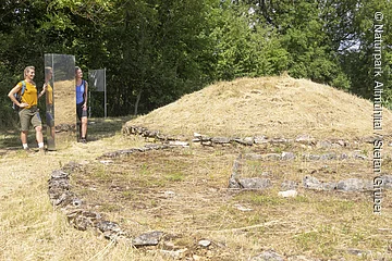 An der Grabhügelanlage stehen zwei Wanderer in sommerlicher Kleidung an einer Stele und bestaunen die Hügelanlage. Hinter ihnen dichter dunkelgrüner Wald.
