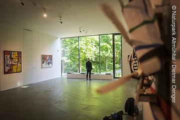 Ein Mann steht vor einer großen hellen Glaswand. Rechts und Links sind Exponate an der Wand zu erkennen.