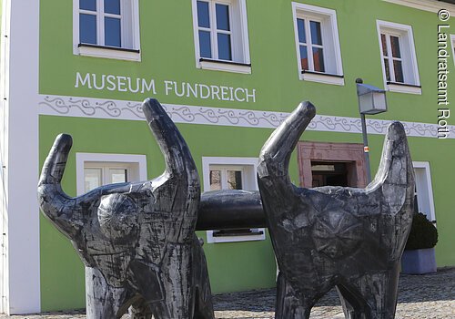 Außenansicht des Archäologischen Museums in Thalmässing. Grünes Haus mit schwarzem Dach. Davor steht eine Skulptur von menschlichen Figuren, die eine Platte tragen.