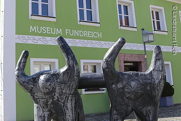Außenansicht des Archäologischen Museums in Thalmässing. Grünes Haus mit schwarzem Dach. Davor steht eine Skulptur von menschlichen Figuren, die eine Platte tragen.