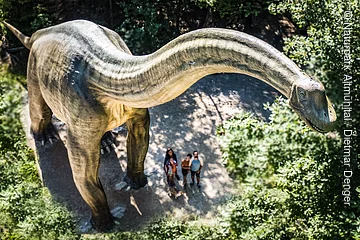 Große Dinosaurierstatue im Wald, Menschen stehen darunter und schauen nach oben.