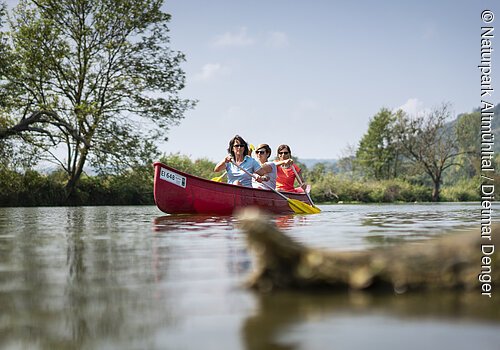 Drei Personen paddeln in einem roten Kanu auf einem ruhigen Fluss mit Bäumen und Hügeln im Hintergrund.