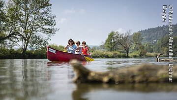 Drei Personen paddeln in einem roten Kanu auf einem ruhigen Fluss mit Bäumen und Hügeln im Hintergrund.