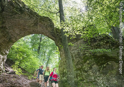 Vier Wanderer gehen einen Waldweg mit Steintreppe unter einem natürlichen Felsbogen hinab.