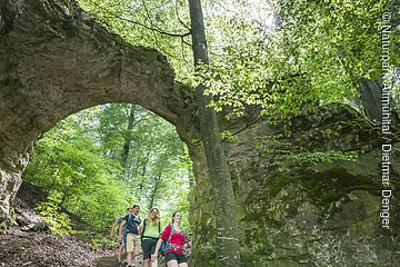 Vier Wanderer gehen einen Waldweg mit Steintreppe unter einem natürlichen Felsbogen hinab.