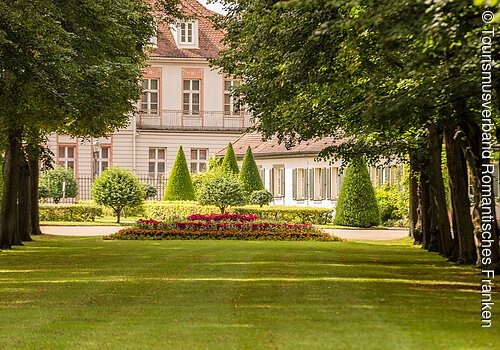 Blick auf einen gepflegten Garten mit Rasen, Bäumen und Blumen vor einem historischen Gebäude mit roten Dächern.