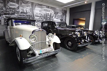 Drei Oldtimer-Autos nebeneinander in einem Museum mit historischem Schwarz-Weiß-Foto im Hintergrund.