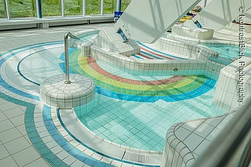 Innenbereich eines Schwimmbads mit kleinem Becken und bunten Fliesen in Regenbogenfarben am Boden.