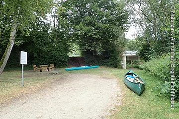 Grüner Kanu und Kajak auf Grasfläche neben Waldweg mit Sitzgruppe und Brücke im Hintergrund.