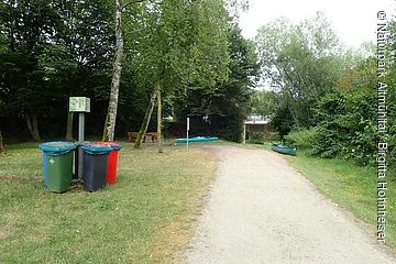 Waldweg mit Mülltonnen, Bänken, Kanus und Brücke im Hintergrund, umgeben von Bäumen und Grasfläche.