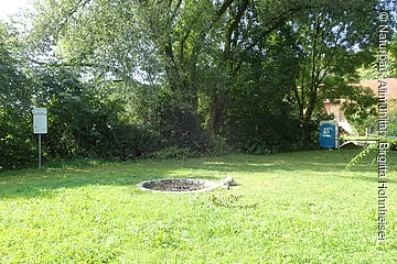 Grüne Wiese mit Feuerstelle, Bäumen, einem Hinweisschild links und einem blauen mobilen Toilettenhäuschen rechts.