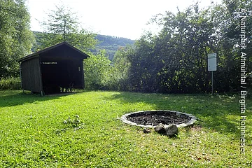 Freistehende Feuerstelle aus Steinen auf einer Wiese neben einer offenen Holzhütte und einem Schild im Waldgebiet