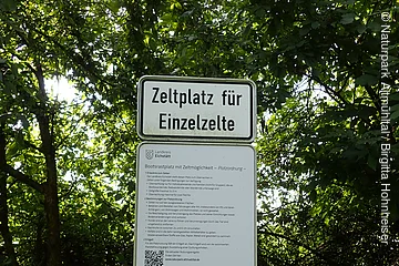Schild mit der Aufschrift „Zeltplatz für Einzelzelte“ und weiteren Informationen vor grünem Laub.