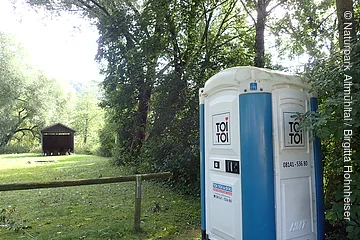 Mobiles Toilettenhäuschen mit blau-weißer Außenwand steht am Waldrand auf einer Wiese neben einem Holzzaun.