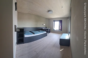 Schlicht eingerichtetes Zimmer mit Einzelbett, Schreibtisch, Stuhl, Fenster und Holzboden.