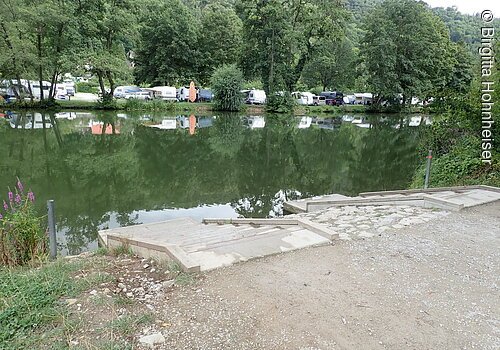 Ufer mit Treppe an einem ruhigen See, gegenüber Bäume und Wohnwagen auf einer Wiese.