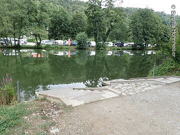 Ufer mit Treppe an einem ruhigen See, gegenüber Bäume und Wohnwagen auf einer Wiese.
