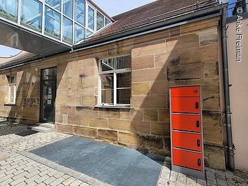 Ein Haus mit brauner Steinfassade. Davor ein Schrnk mit roten Schließfächern. Zum Dach des einstöckigen Hauses führt ein verglaster Gang.